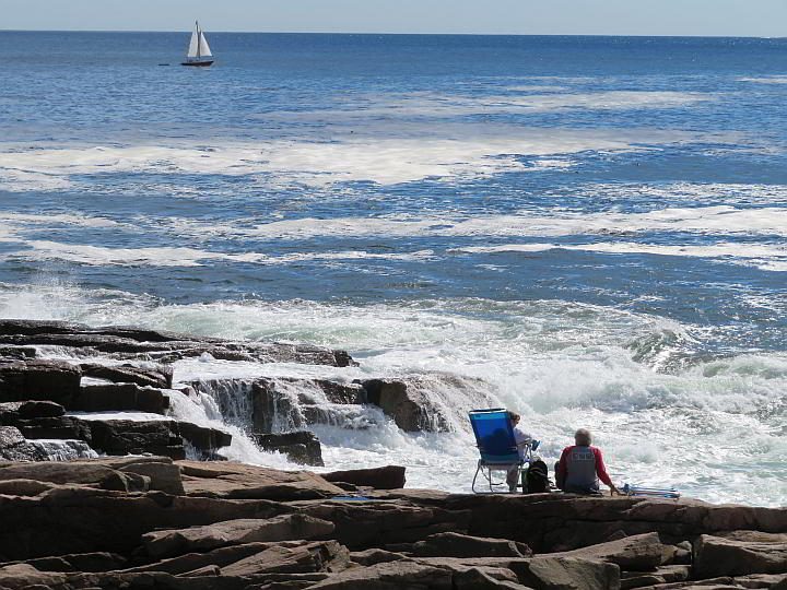 在阿卡迪亚国家公园，两名游客坐在岩石海岸线上观看海浪和帆船
